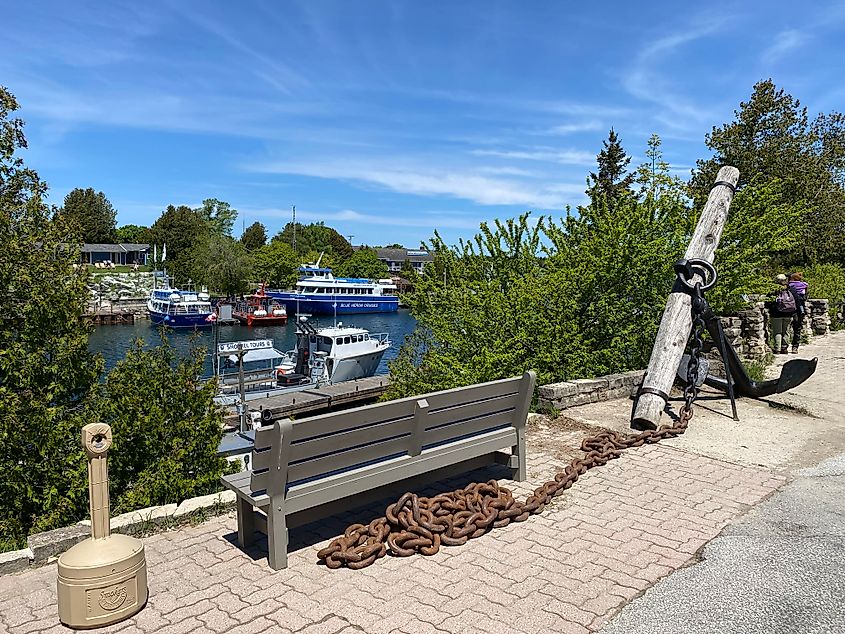 A large black ship anchor with rusty chain sits next to a bench overlooking a small marina