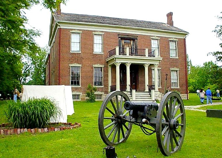 A historic red-brick house with white trim stands in a green lawn. A cannon and a white tent are in the foreground, conveying a historical atmosphere.