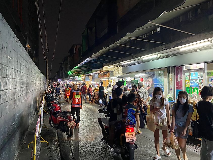 Night on a crowded side street in Bangkok lined with motorcycles on the left, shops/food carts on the right.