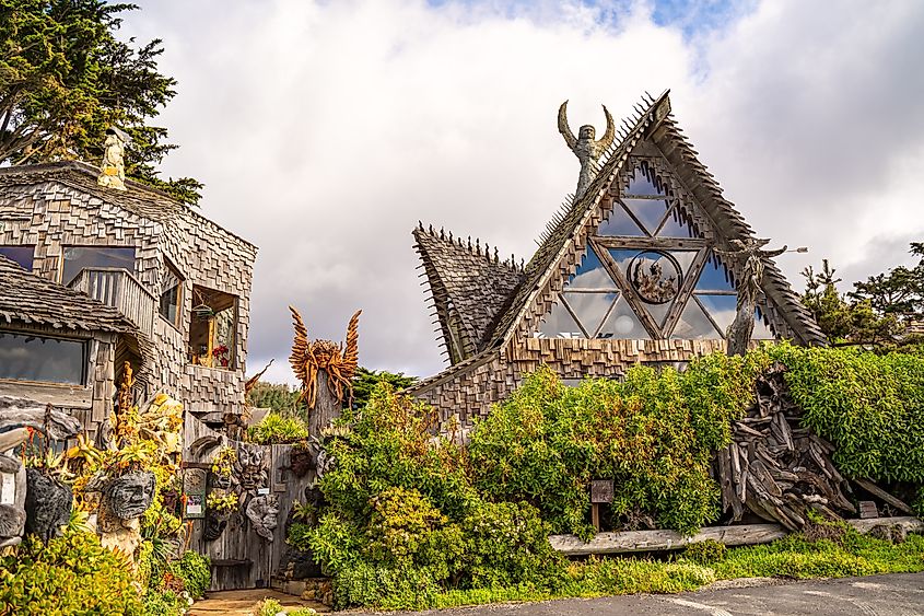 Unique house with wooden shingles and angular roofs, adorned with sculptures and surrounded by greenery. An overcast sky adds a mysterious tone.