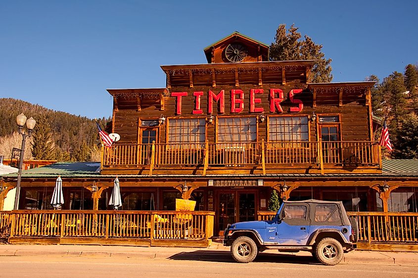 Rustic wooden restaurant with a red "Timbers" sign, American flags, a balcony, and a blue Jeep parked in front. Mountain forest backdrop under clear sky.