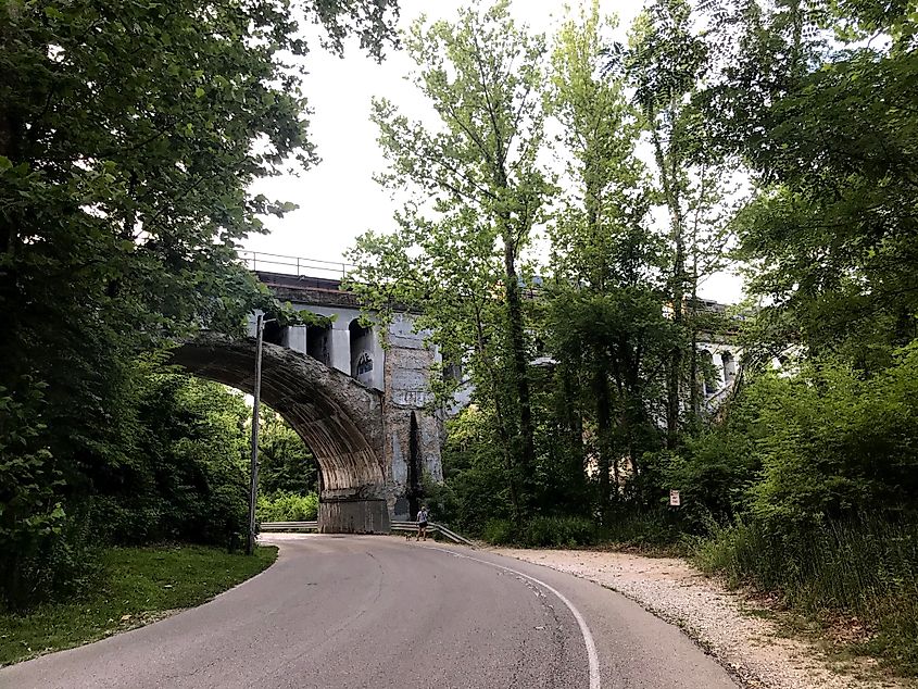 Curved road passing under a tall, stone bridge surrounded by lush green trees. Soft light filters through, creating a serene, peaceful atmosphere.