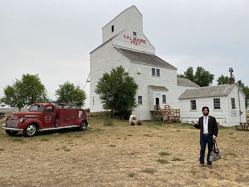 A man dressed in old ranching attire stands outside of a classic prairie grain elevator, now a local museum. 