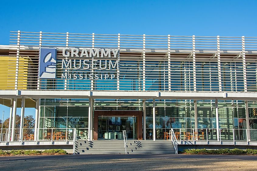 Front view of the GRAMMY Museum Mississippi building, featuring a modern glass façade with horizontal white slats and a large sign displaying the museum's name. Steps lead up to the entrance, with outdoor seating visible on both sides.