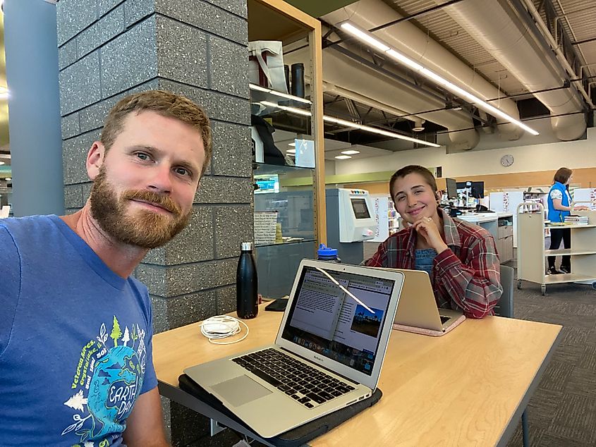 A couple sits at a library table, both posing for a picture before resuming work at their laptops.  