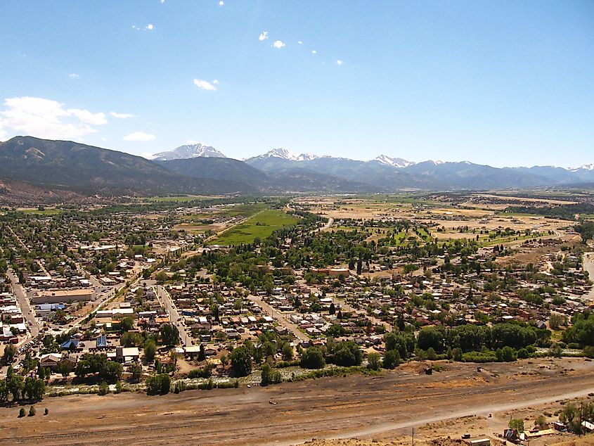 Aerial view of Salida, Colorado, showing a small town with houses, streets, and green fields in a broad valley, with snow-capped mountains in the distance under a clear blue sky.