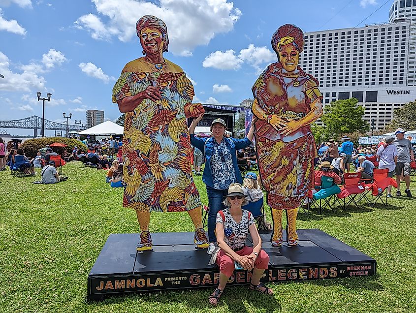 Visitors at the JAMNOLA Garden Legends exhibit in New Orleans, Louisiana, posing with life-sized cutouts of cultural figures. The background features the Mississippi River, a bridge, and a crowd enjoying the outdoor event.