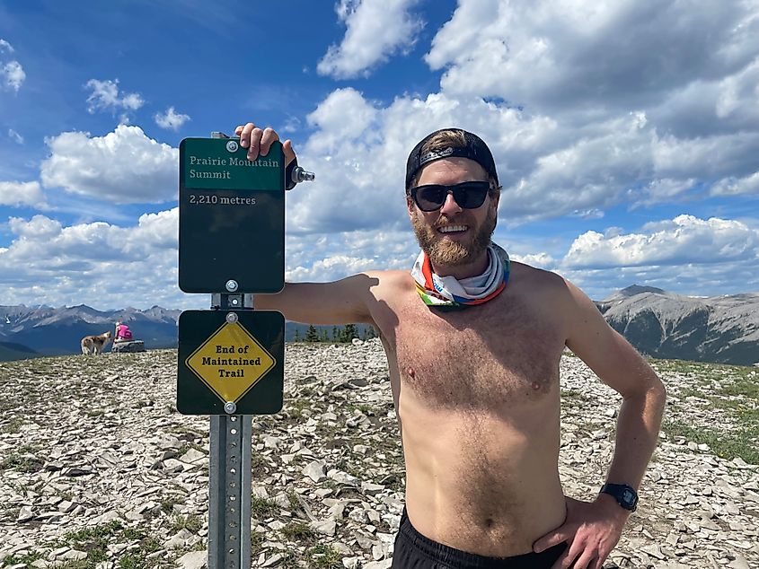 A shirtless male runner poses beside the Prairie Mountain summit marker on a fine, partly cloudy day. 