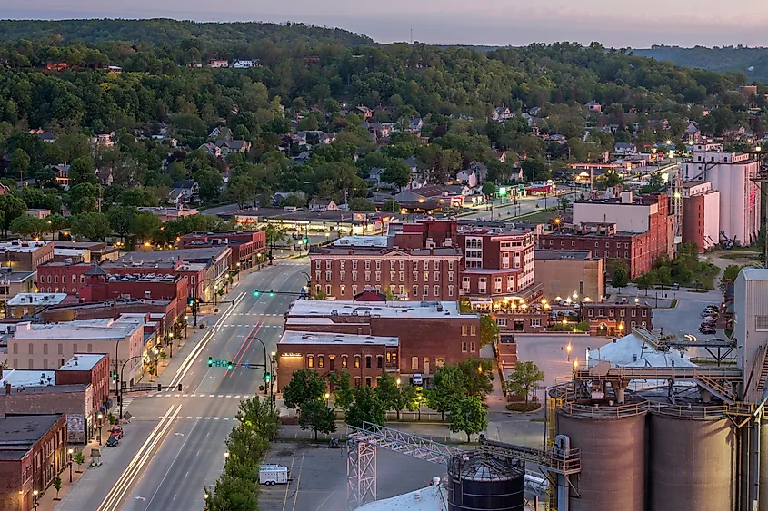 Aerial view of a small town at dusk, featuring illuminated streets lined with brick buildings. Green hills rise in the background under a pink sky.