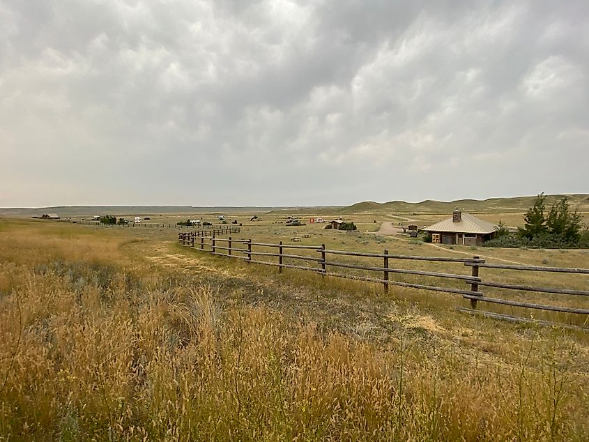 A prairie campground as seen from the other side of a basic wooden fence. One central building acts as the focal point, the rest is spacious RV and tent sites. 