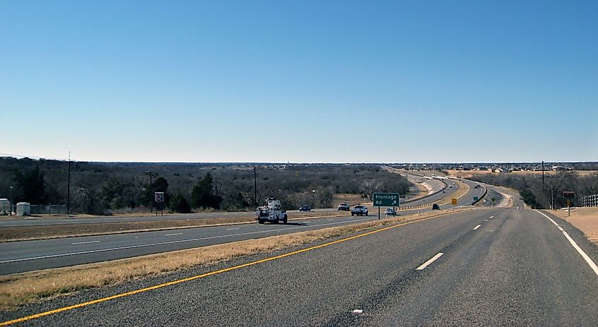 U.S. Route 84 in Woodway, Texas, showing the exit onto a frontage road, surrounded by grassy shoulders, road signs, and light traffic under a clear sky.