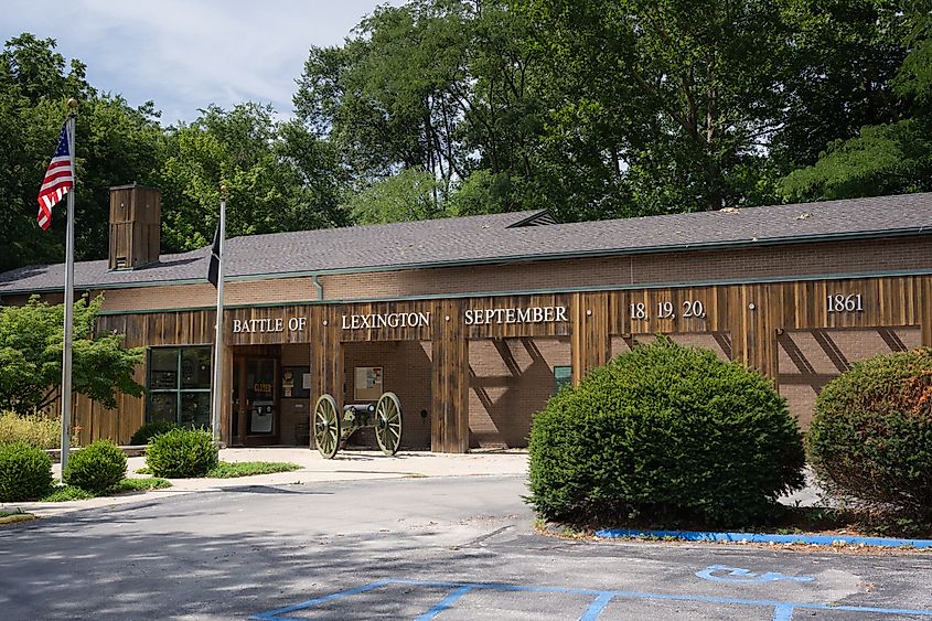 The image shows the Battle of Lexington State Historic Site entrance with a wooden facade, vintage cannon, and American flag, surrounded by greenery.