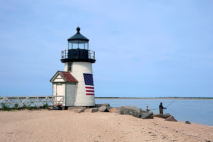 Small lighthouse on a sandy shore in Nantucket, Massachusetts with an American flag displayed on its side, as two people fish on nearby rocks by calm water.