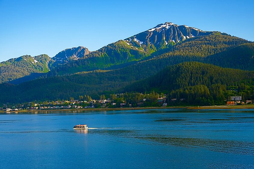 A scenic view of Juneau, Alaska, with a boat gliding across calm blue waters, backed by a lush forested shoreline and towering snow-capped mountains under a clear blue sky.