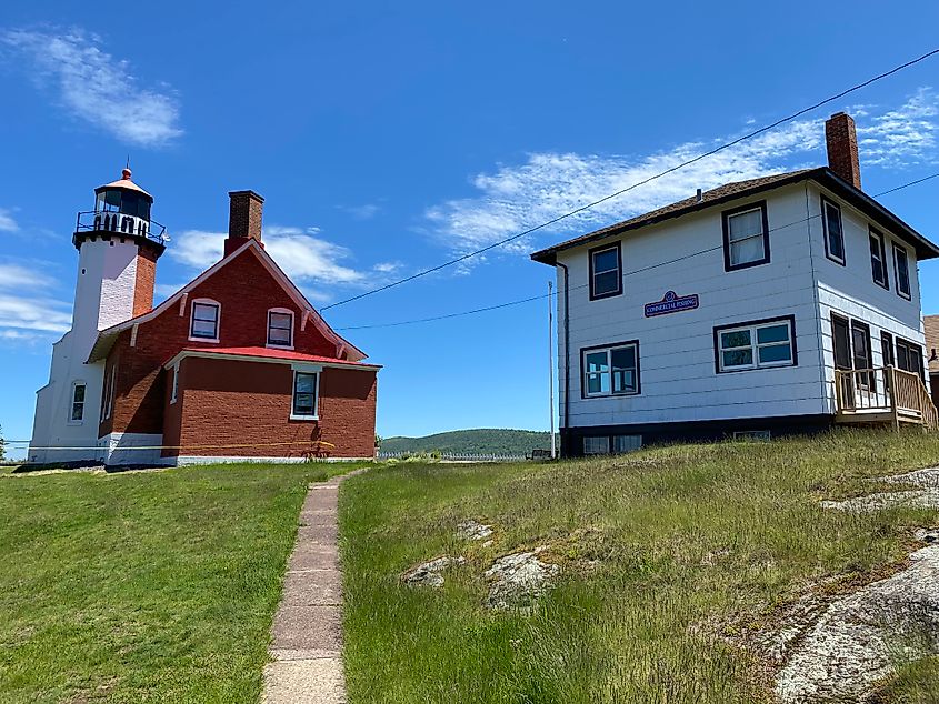 A red brick lighthouse stands next to a boxy white commercial fishing structure atop a grassy hill.