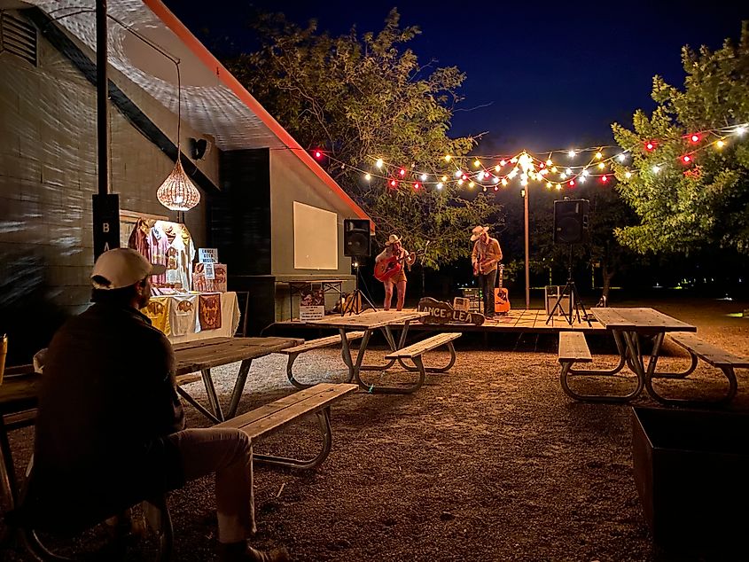 A male and female musician entertain one man sitting at a collection of picnic tables. It’s nighttime and colourful lighting hangs over the small wooden stage. 