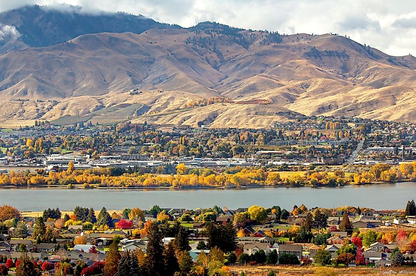 A panoramic view of a town in autumn, with colorful foliage in the foreground, a calm river, and rolling hills under a partly cloudy sky in the background.