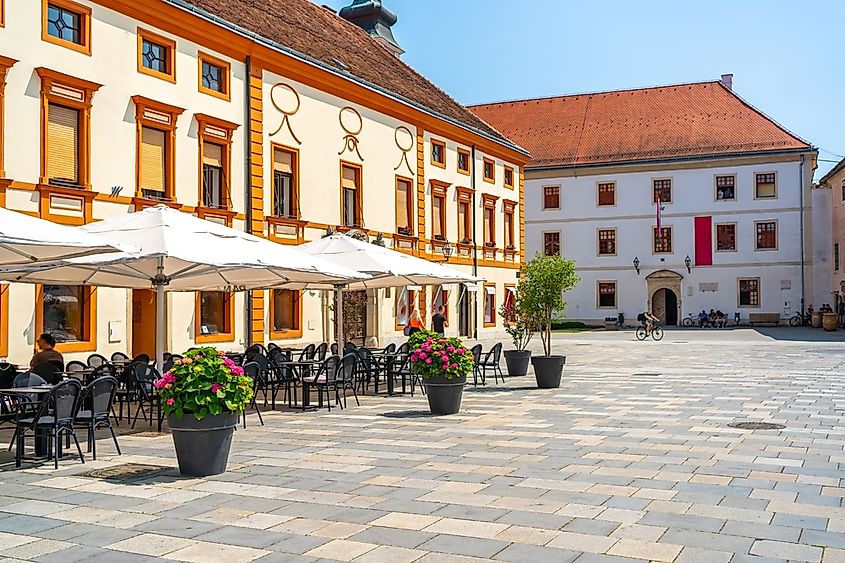 Vibrant town square in Varaždin with yellow-orange facades, outdoor seating, and people enjoying a sunny day in a lively atmosphere