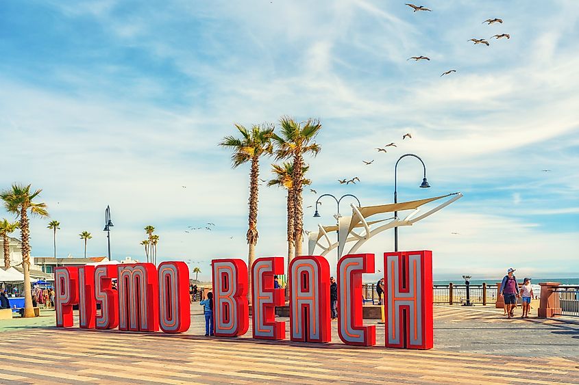 Large red letters spell "Pismo Beach" in front of palm trees and a boardwalk under a blue sky with scattered clouds and flying seagulls.