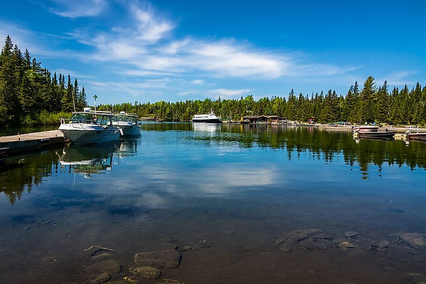 Boats rest on calm water at Rock Harbor in Isle Royale National Park, with a forested shoreline and blue sky reflected on the surface of Lake Superior.