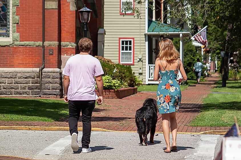 A young couple with their dog strolls through the historic district of Chestertown, Maryland. The woman, in a floral summer dress, has light hair, while the man wears a t-shirt and jeans.