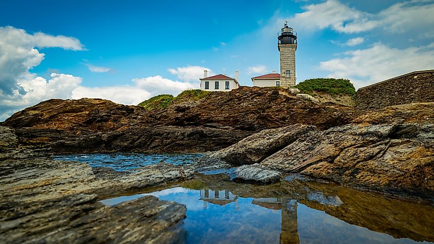 Beavertail Lighthouse in Jamestown, Rhode Island, with a dramatic cloudscape reflecting over the water and rugged rocky cliffs in the foreground.