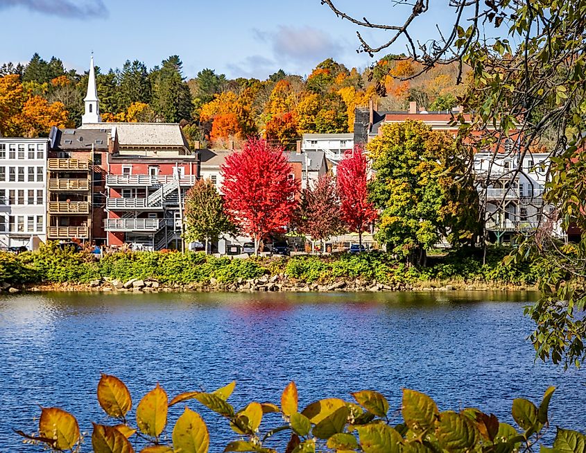 Quaint town by a lake in autumn. Vibrant red and orange trees line the water, with rustic buildings and a church spire in the background. Calm and picturesque.