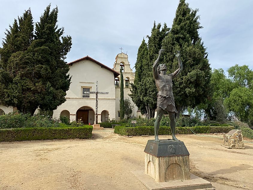 A statue with a famished man raises his hands to the heavens stands out front of an old Spanish mission.