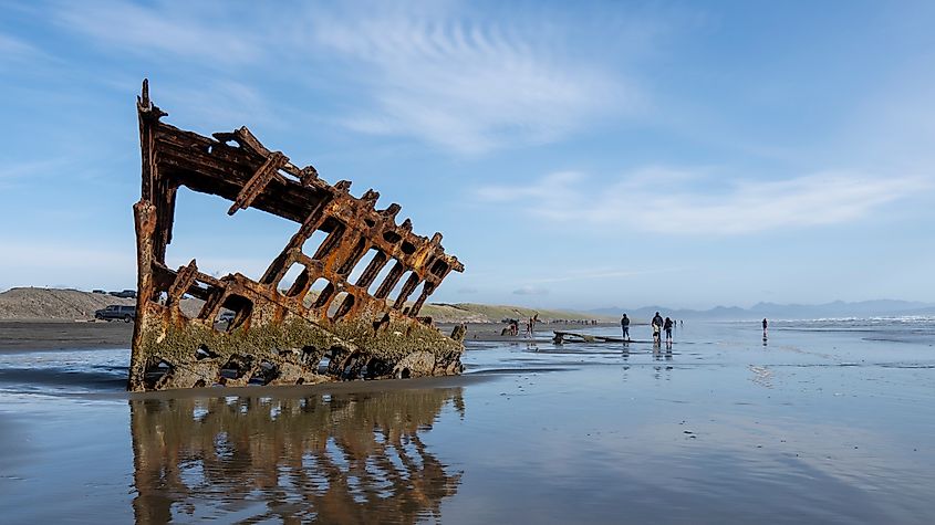 Rusty shipwreck remains partially buried in wet sand on a wide beach under a clear blue sky. People in the distance explore the shoreline.