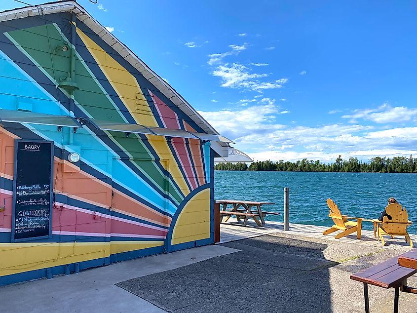 Someone sits in a set of lakeside yellow cottage chairs beside technicolored bakery. 