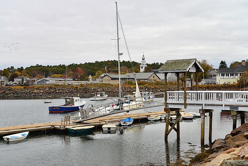 Harbor scene with small boats docked at a wooden pier, a sailboat nearby, and a quaint town with a white church and autumn trees in the background. Calm and serene ambiance.