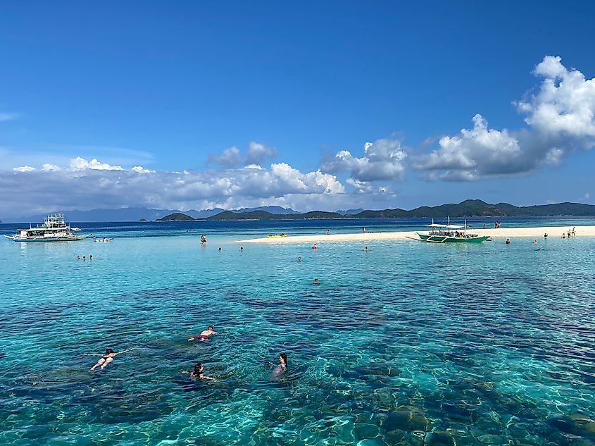 Small boats anchor near a sandy island while occupants snorkel amongst the crystal clear waters.
