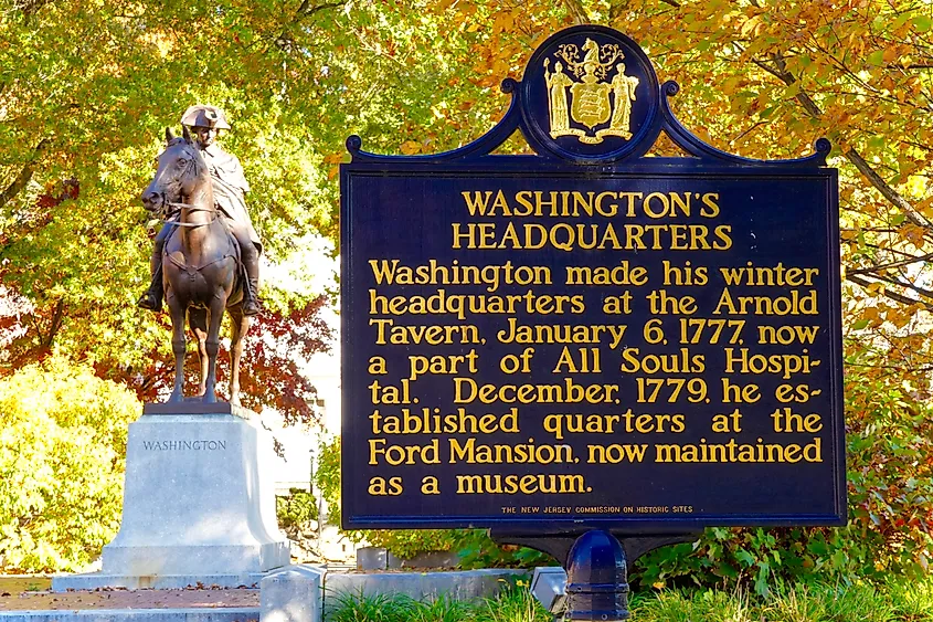 A statue of a historical figure on horseback stands beside a large informational sign about Washington's Headquarters, set against autumn foliage.