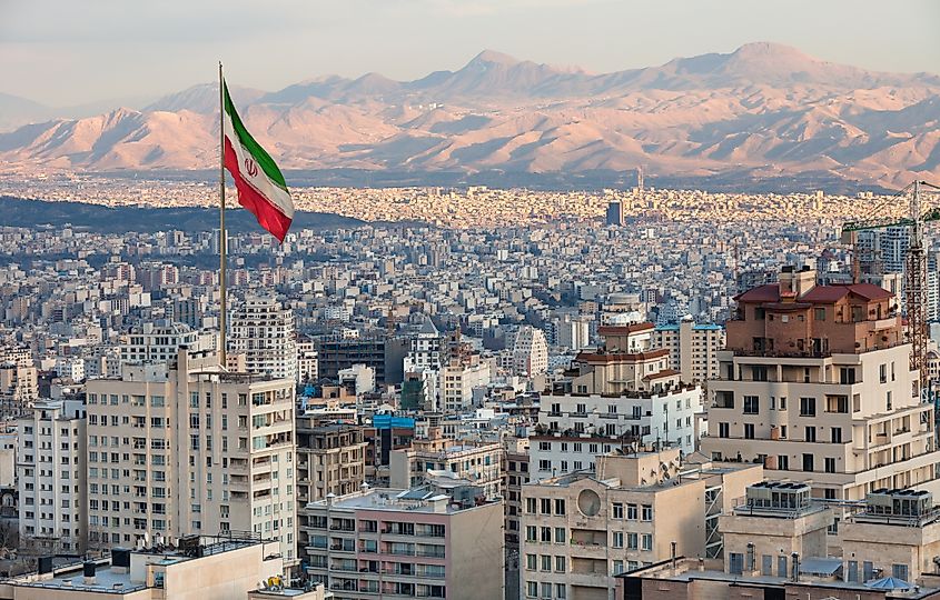 Waving Iran flag above skyline of Tehran at sunset.