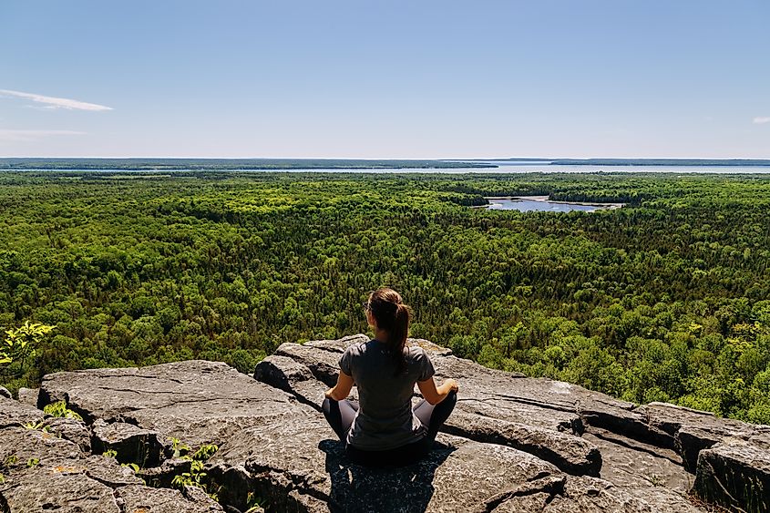 A woman sits cross-legged atop a cliff, looking out over an expansive forest