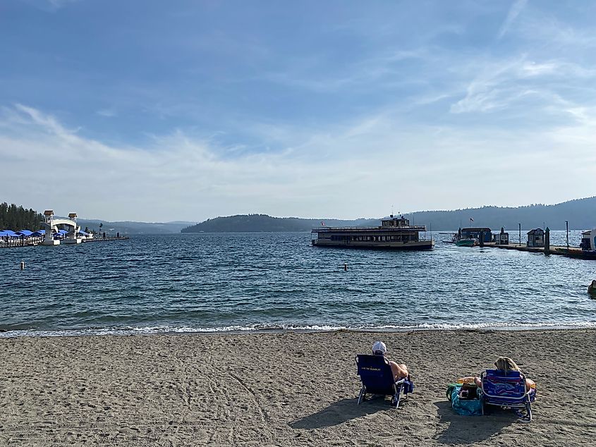 A couple in beach chairs enjoys a small sandy lake beach. A tourist cruise boat can be seen parking at the lengthy dock.