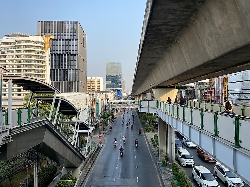 A main roadway in Bangkok as seen from the Skytrain platform above. Pedestrians cross the elevated skywalk, motorcyclists move freely in the left lane, while cars are jammed up on the right.