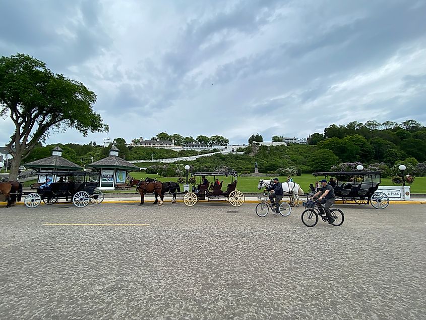 A couple of cyclists share the road with horse-drawn carriages. Fort Mackinac can be seen on the green hill above 