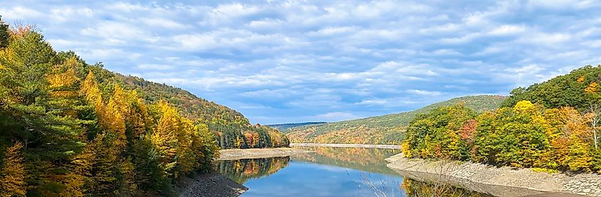 Calm lake surrounded by forested hills with vibrant autumn leaf colors, reflecting in the water with low water levels, under a blue sky with clouds. Pepacton Reservoir, New York State.