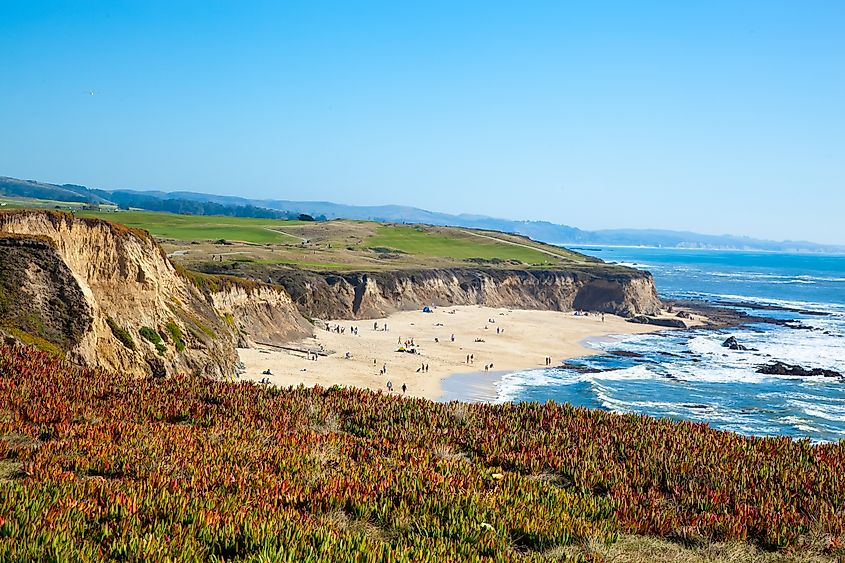 Cliffs with green and red vegetation overlook a sandy beach with small groups of people. The ocean waves gently crash under a clear blue sky.