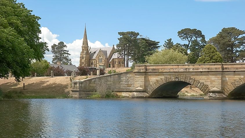 A close-up view of the historic stone bridge and church in Ross, Tasmania, Australia. The bridge’s detailed stonework spans a calm river, with the church standing in the background, embodying the town’s colonial charm.