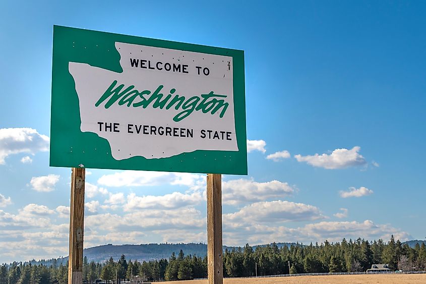 A roadside sign with welcome to Washington State on it, with a rural setting and blue sky behind.