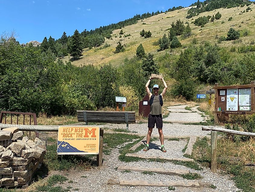 A male hiker holds a rock over his head at the trailhead of the M-Trail in Bozeman, Montana.