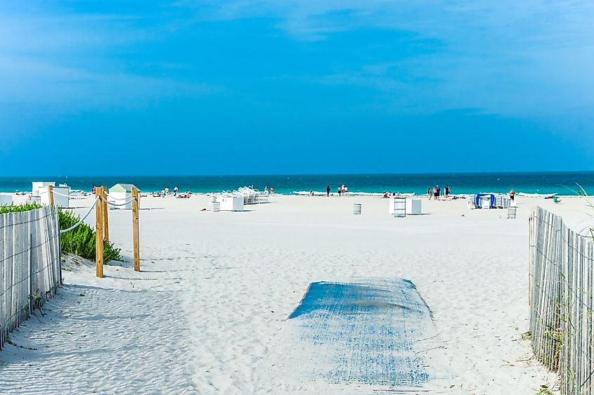 A sunny day at the beach in Gulf Shores, Alabama, with a pathway leading to the sandy shore and the ocean in the background. People are enjoying the beach, lounging, and swimming under a clear blue sky.