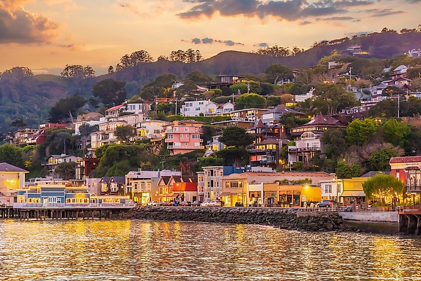 Charming hillside town at sunset, featuring colorful houses atop lush greenery, with reflections of warm lights shimmering on calm waterfront. Serene ambiance.