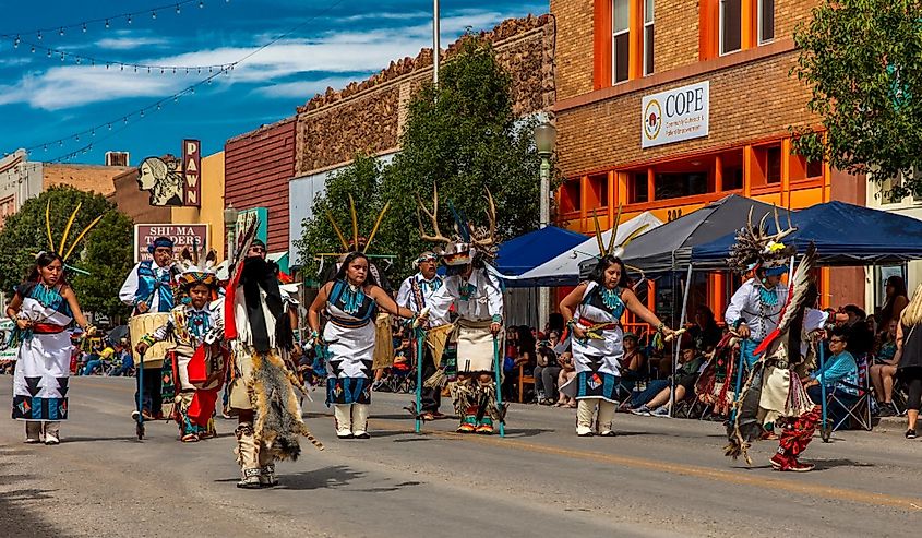A vibrant parade with people in traditional attire featuring antler headdresses and decorative elements dances down a street. Spectators watch from the sidewalk.