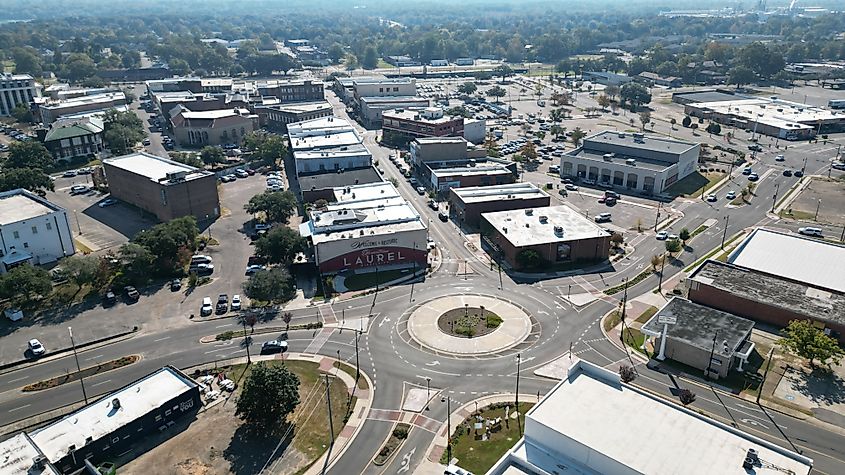 Aerial view of a small downtown area in Laurel, Mississippi, centered around a roundabout intersection. Low-rise commercial buildings, parking lots, and tree-lined streets surround the circle, with light traffic and a large mural reading 'Laurel' visible on one building.