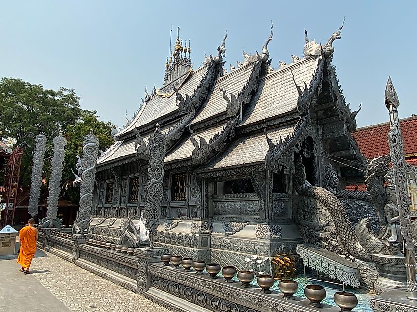 An ordained monk in orange robe walks beside an impressive silver temple in the bright mid-day sun.