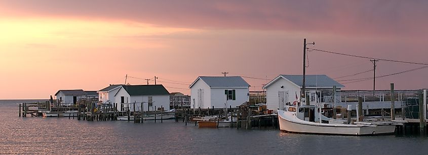 Dawn breaks over Tangier Island, Virginia, casting a soft pink glow on the sky and reflecting on the calm waters, with wooden fishing shacks, docks, and boats lining the shoreline.