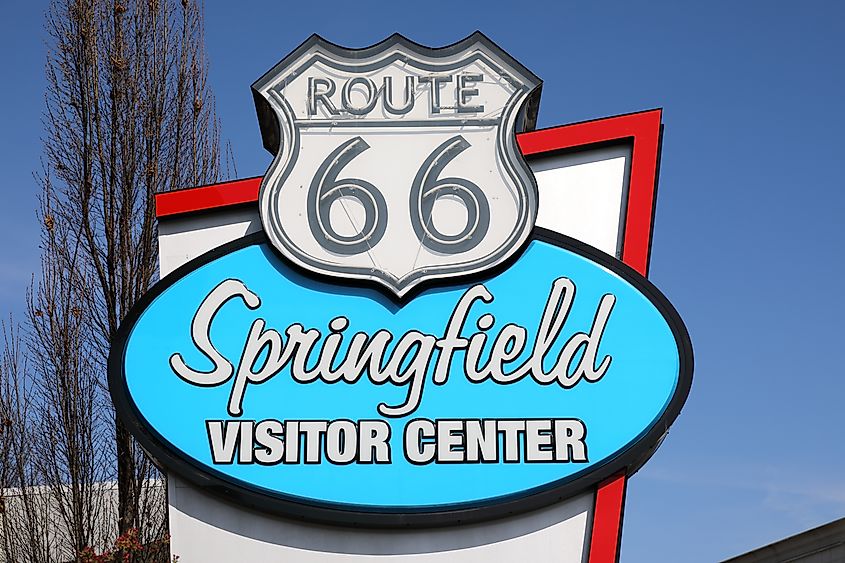 A Route 66 sign with "Springfield Visitor Center" in bright blue and white lettering. Tall trees and a clear blue sky are in the background.